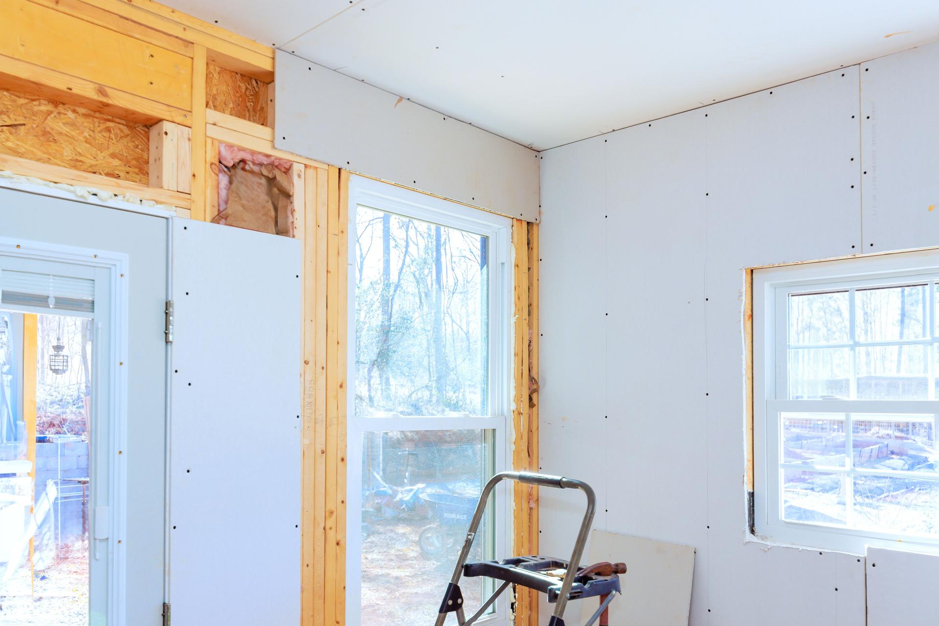 Drywall installation process in a bright, unfinished room with large windows during daytime