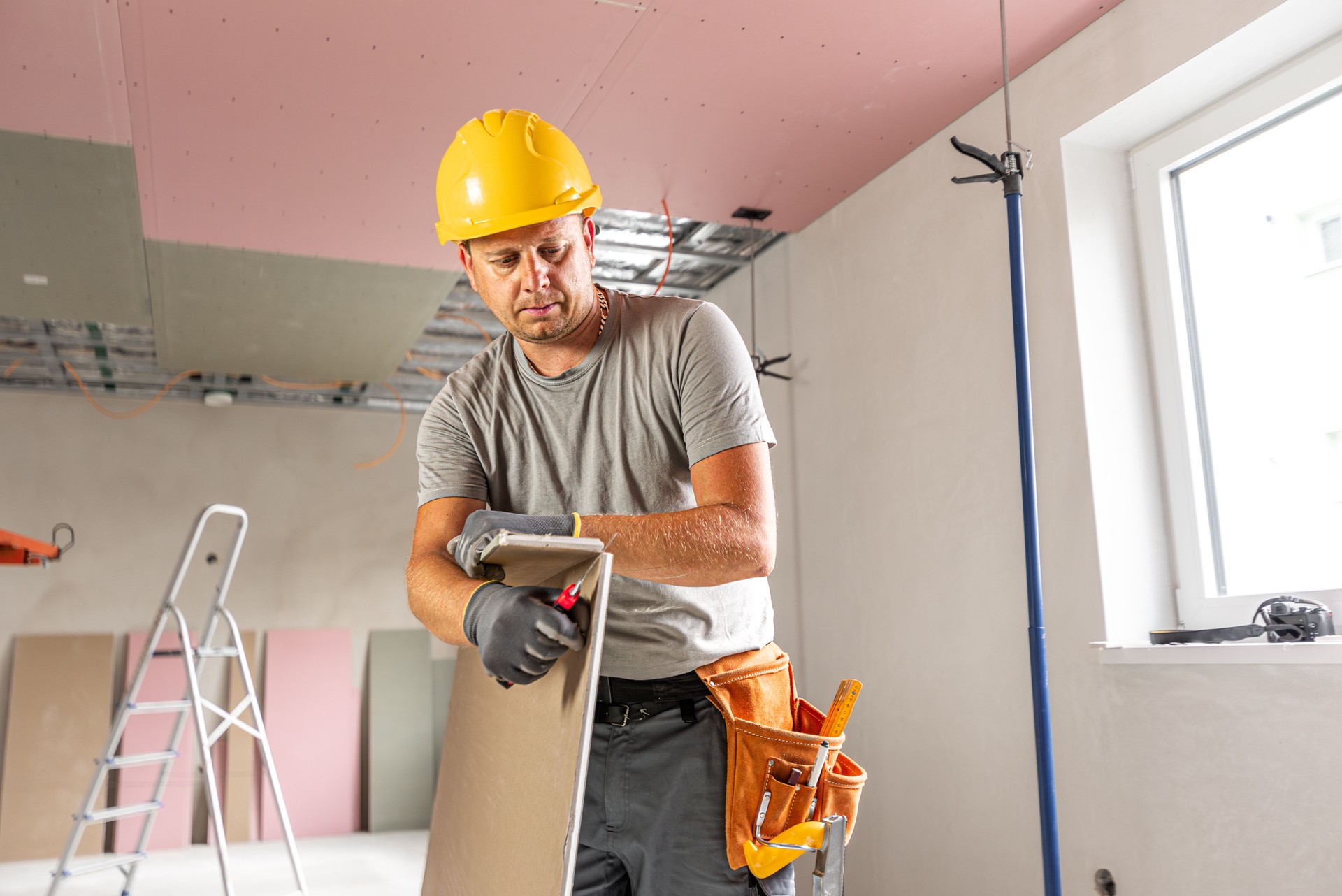 The worker make a plasterboard ceiling. He doing adjust a size of the board using knife cutter. A red plasterboard improve the fire resistance of ceiling structures.